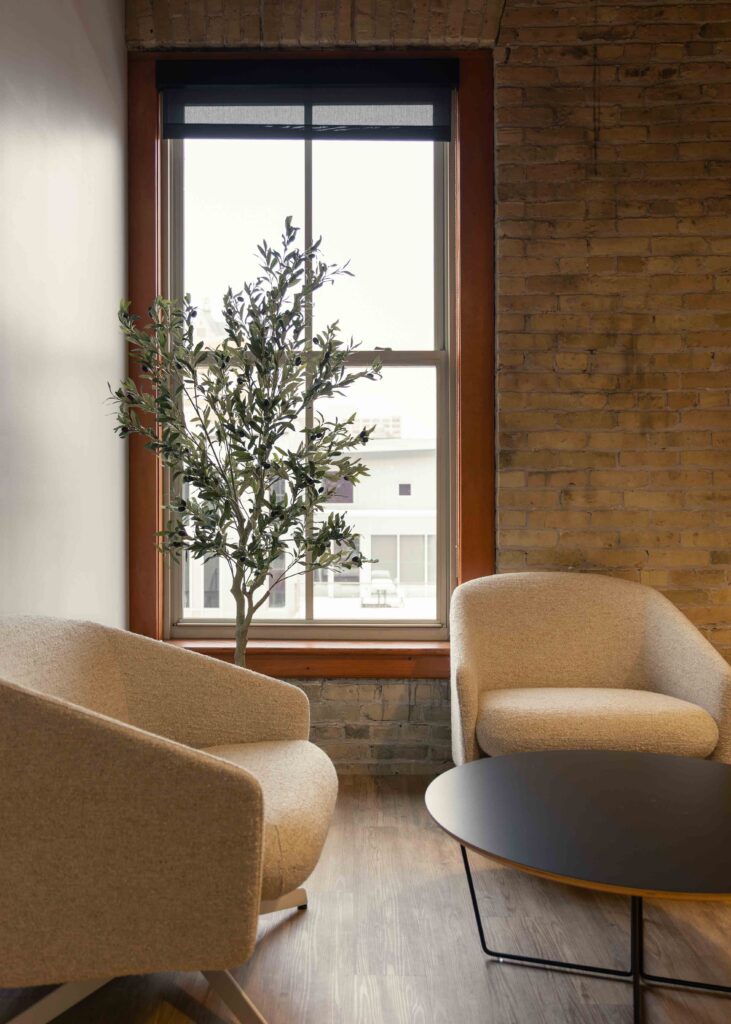 Small lounge area in Branigan’s Milwaukee office with two upholstered armchairs, a round coffee table, and a potted tree in front of a tall window and exposed brick wall.