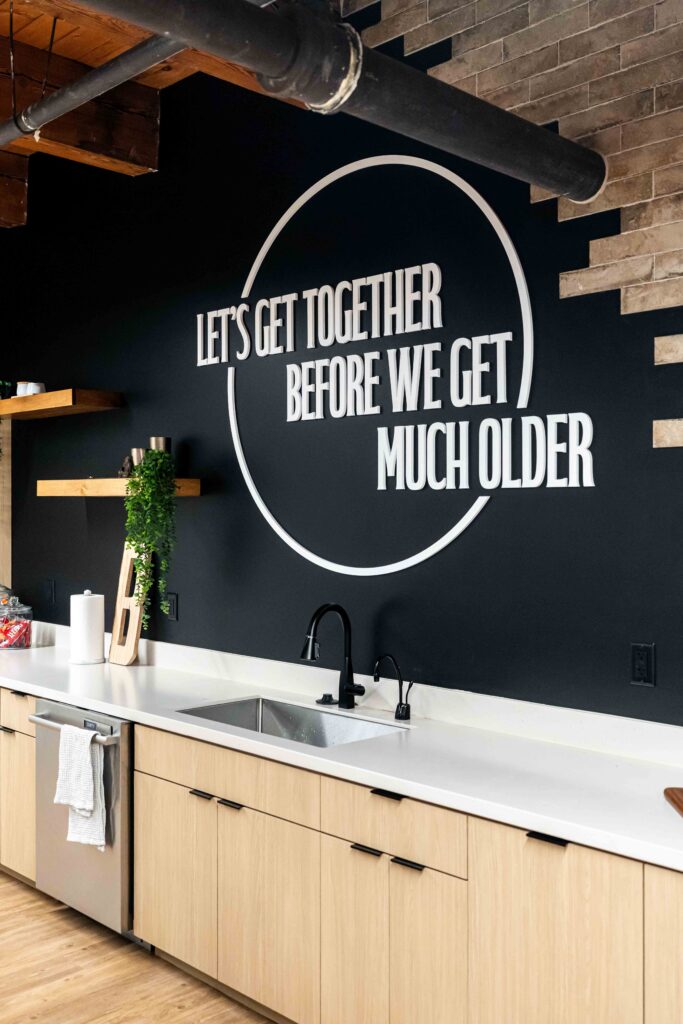 Breakroom in Branigan's Milwaukee office featuring a black accent wall with the phrase, "Let's get together before we get much older," in white lettering above light wood cabinets and shelving