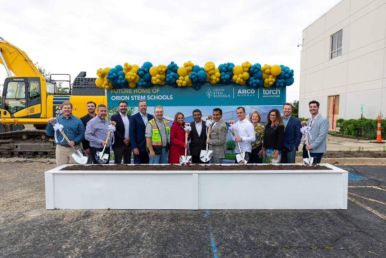 Group of project partners holding ceremonial shovels at the Orion STEM Schools groundbreaking, standing in front of a site sign with a balloon arch and excavator behind them.