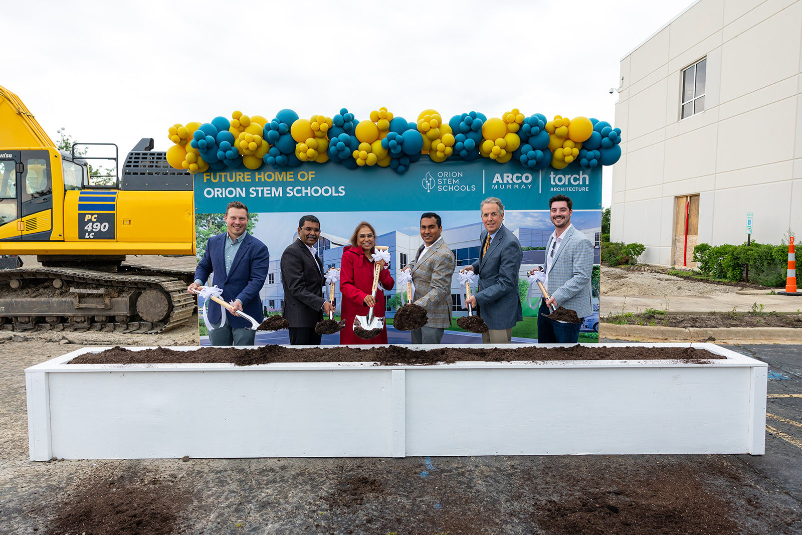 Six project partners ceremonially toss soil with shovels at the Orion STEM Schools groundbreaking, standing in front of a campus sign with a balloon arch and excavator behind them.