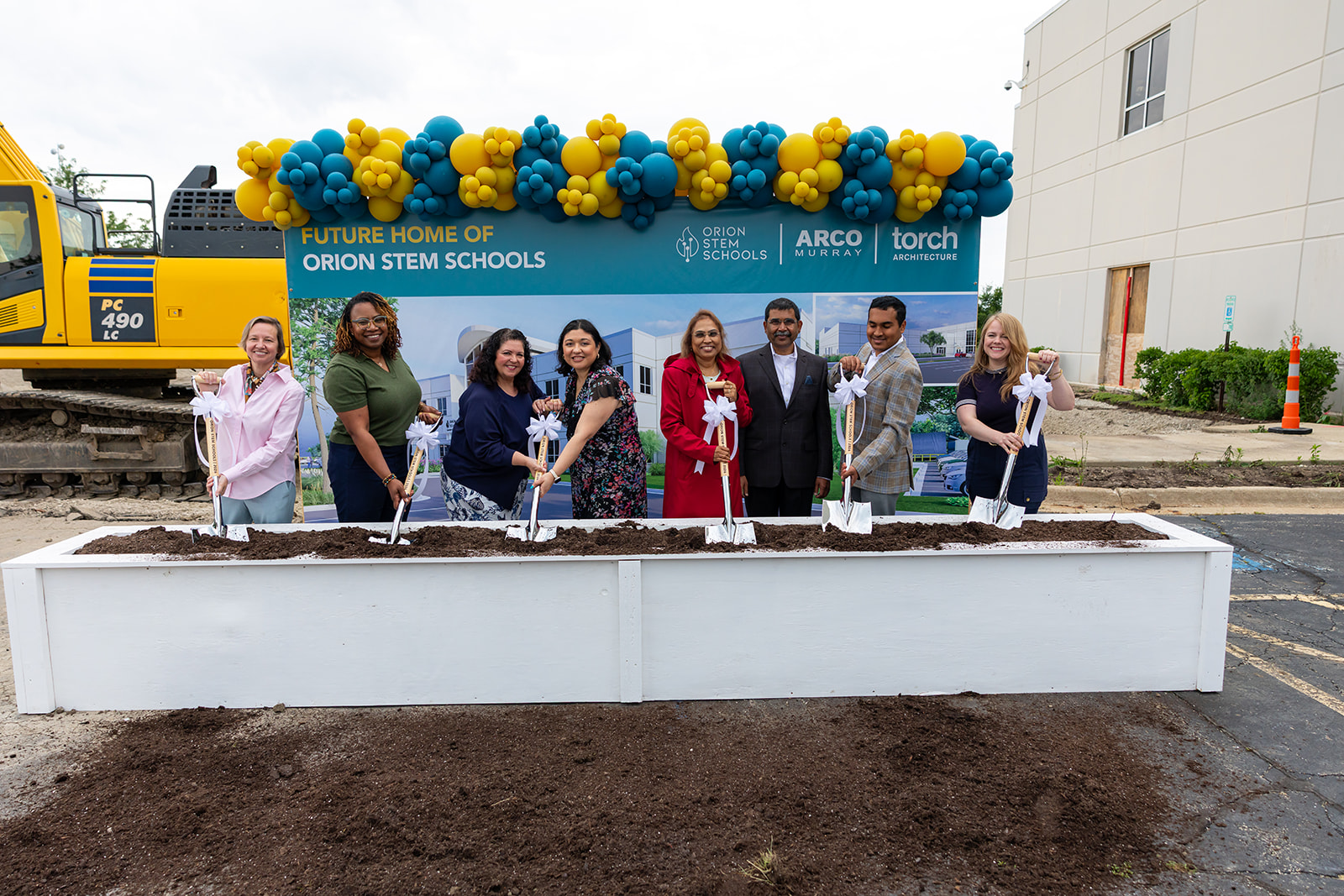 Orion STEM Schools staff and partners pose with ceremonial shovels at the groundbreaking, standing in front of the future campus sign with a balloon arch and excavator behind them.