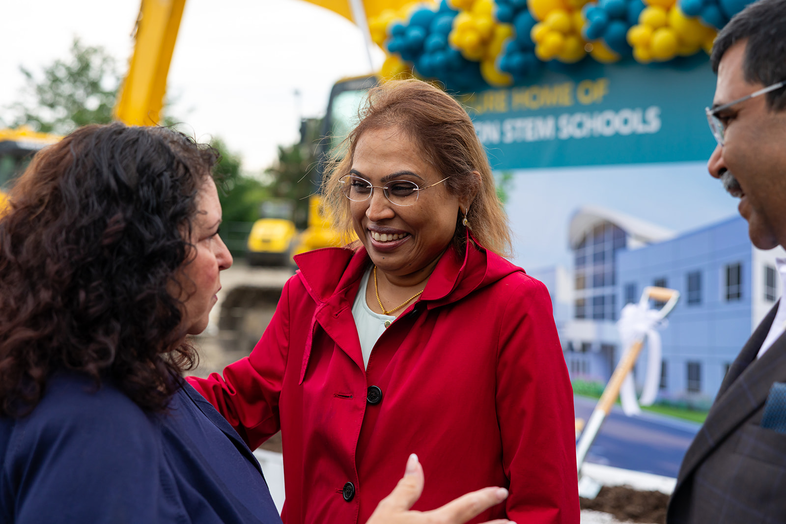 Selvei Rajkumar speaks with two attendees at the Orion STEM Schools groundbreaking, with construction equipment and a project rendering in the background.