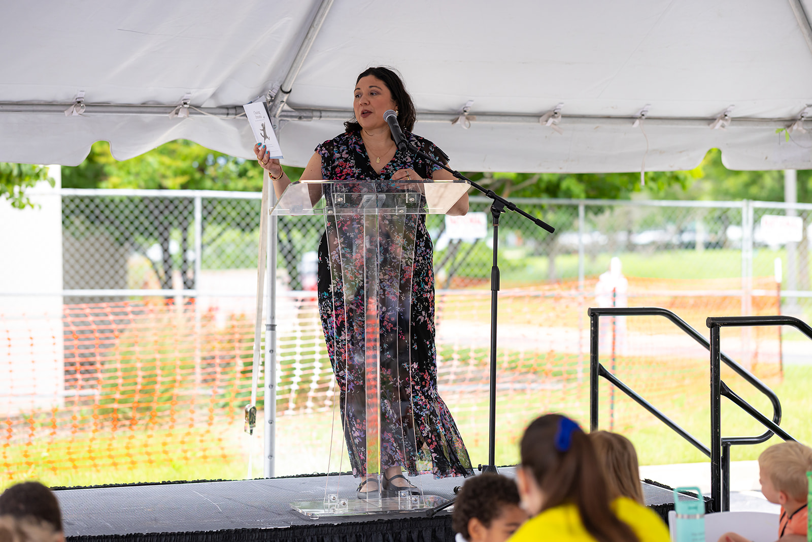 A woman reads a storybook aloud to children from a podium under a tent at the Orion STEM Schools groundbreaking event.