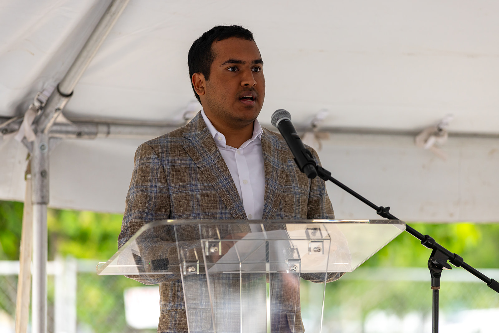 A man delivers remarks from a clear podium under a tent at the Orion STEM Schools groundbreaking event.
