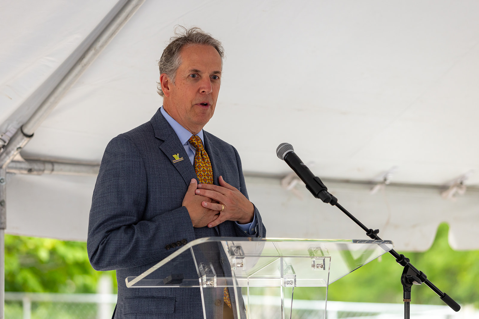 Mayor of Warrenville, Andrew Johnson, speaks at a clear podium under a tent during the Orion STEM Schools groundbreaking event.