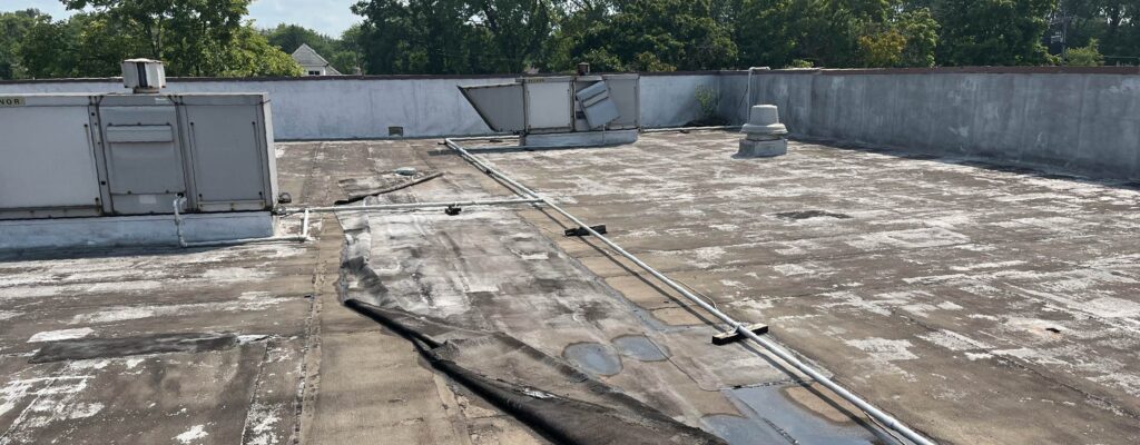 Chicago Collegiate Charter School's weathered rooftop before renovations. Peeling roofing material, exposed seams, HVAC units, pipes, and a drain; trees and homes are visible beyond the parapet.