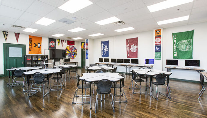 Classroom with rows of tables and chairs, wall-mounted computer stations, school pennants, and polished wood floors.