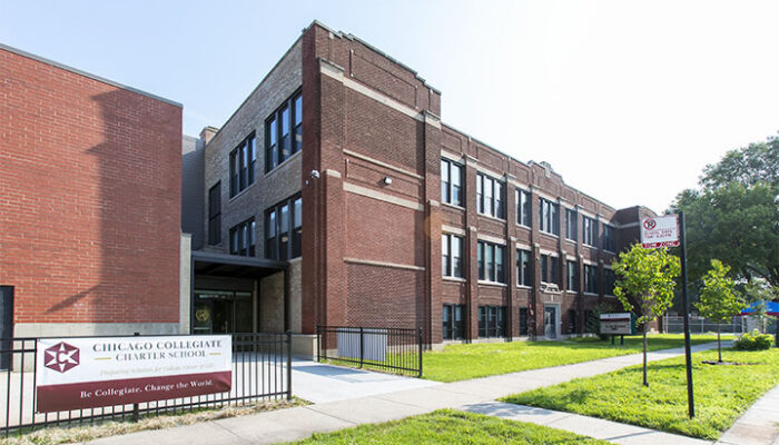 Red-brick, three-story school building with a front lawn, sidewalk, and school sign in the foreground.