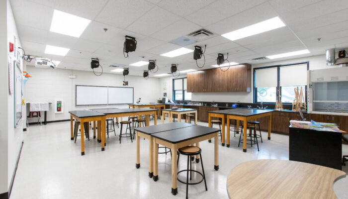 Science lab with black-topped workstations, sinks and faucets, stools, wall-mounted display screens, and wood cabinets.